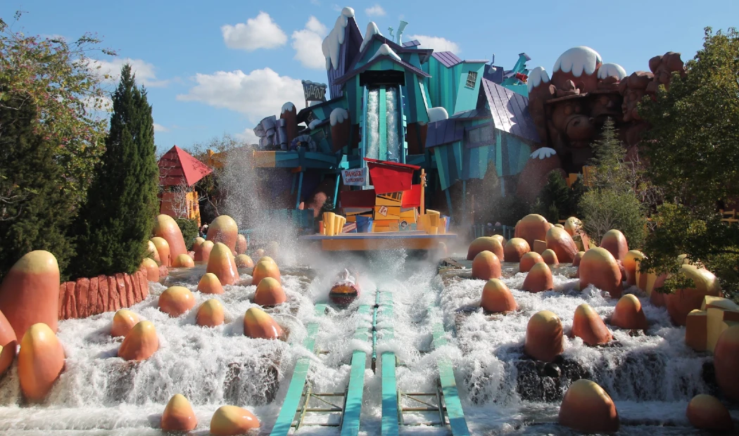 A colorful amusement park water ride splashes down a track surrounded by oversized cartoonish foliage and rock formations
