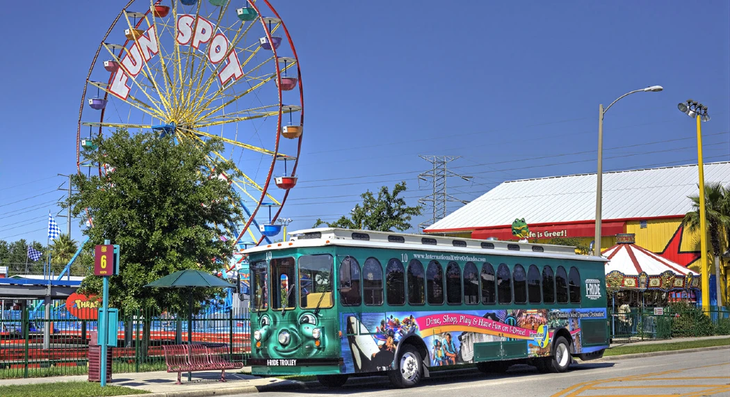 A green I-RIDE trolley bus parked near a colorful ferris wheel and amusement park
