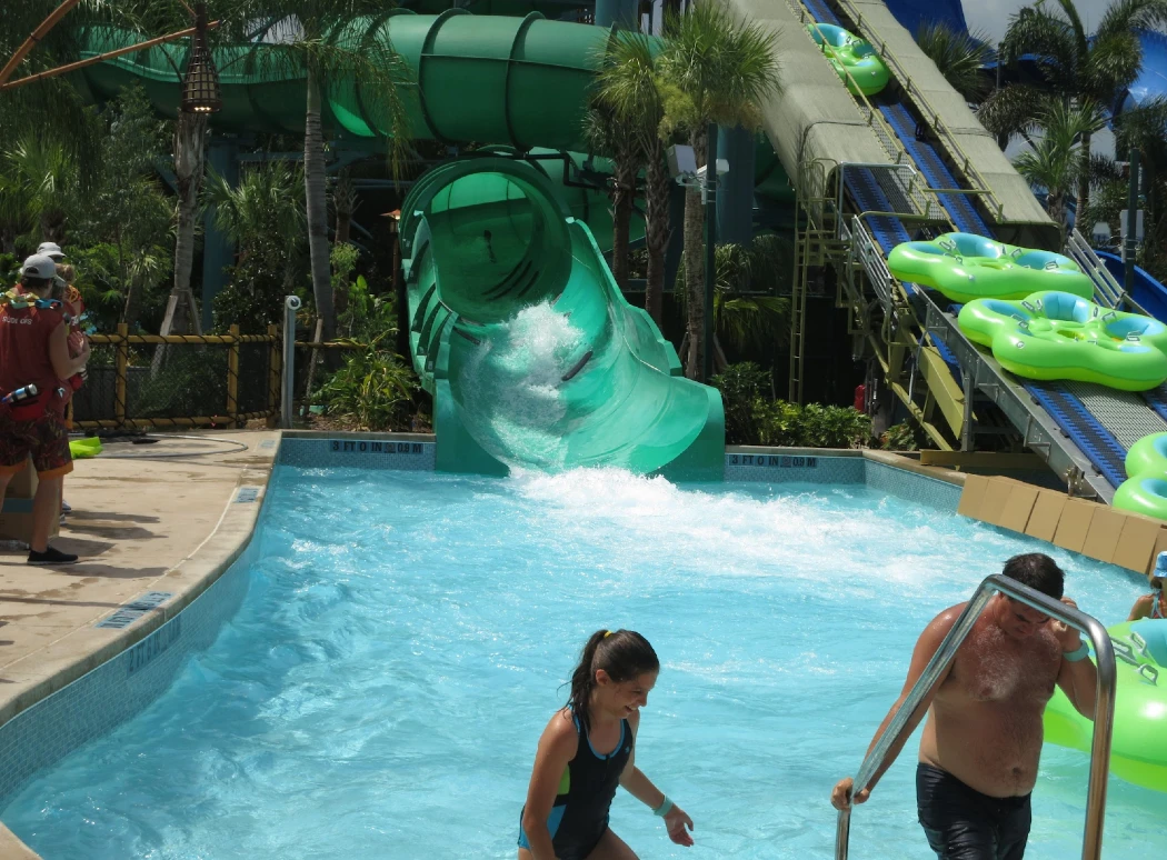 A water park scene featuring a tall, green waterslide, guests enjoying the pool, and inner tubes awaiting riders