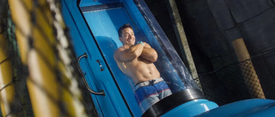 A muscular man poses inside a blue aquatic capsule while water streams down the glass