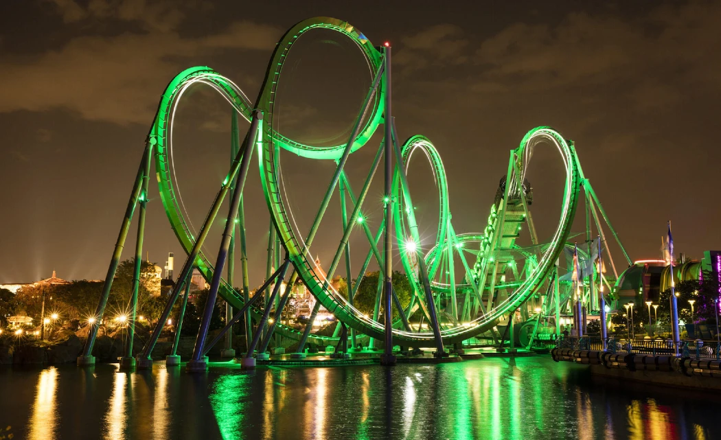 Illuminated Incredible Hulk Coaster coaster at night, featuring green lights, twisting loops, and reflections on the water below