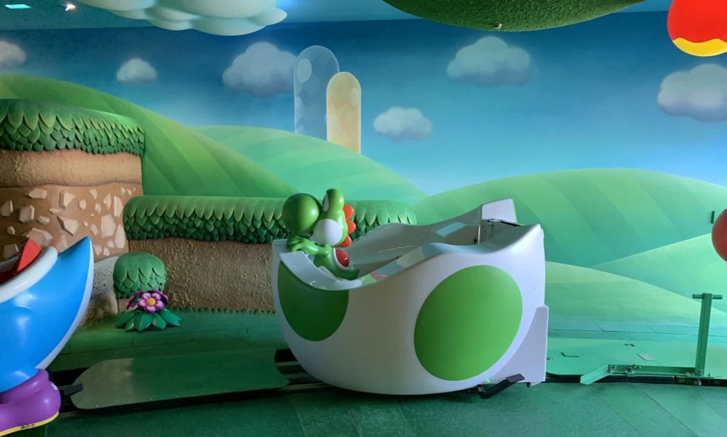 A colorful indoor amusement ride themed after Yoshi, set against a vibrant landscape of green hills and cartoonish clouds