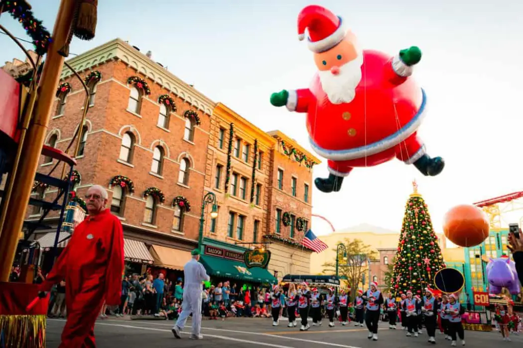 A giant inflatable Santa Claus hovers over a festive parade, with a decorated Christmas tree and performers in holiday attire marching below