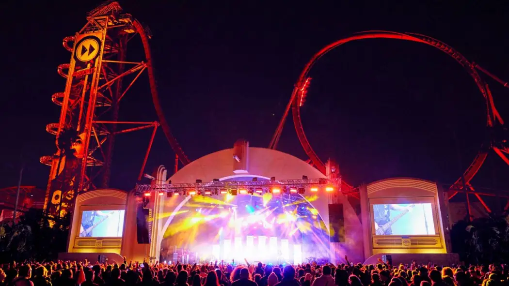 A vibrant concert scene at night with a large crowd, illuminated by colorful lights, Set against a backdrop of a towering roller coaster
