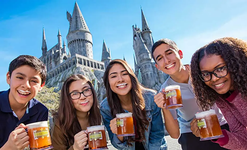 Group of friends holding drinks in front of a grand castle on a sunny day, showcasing a theme park atmosphere