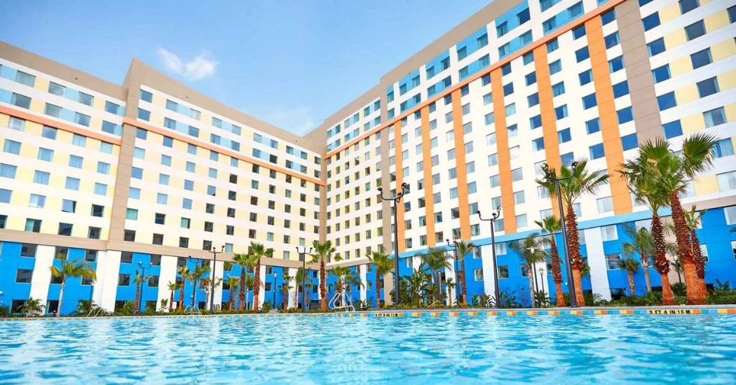 Sunlit hotel with colorful architecture, framed by palm trees, alongside a sparkling swimming pool under a clear blue sky