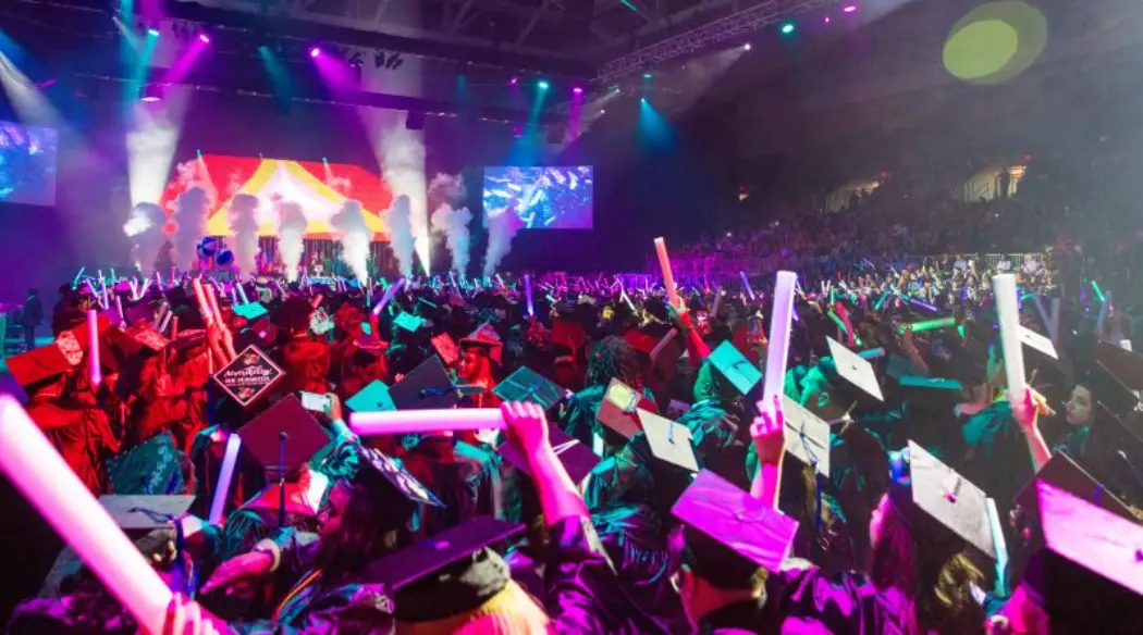 A vibrant graduation ceremony with graduates in caps and gowns waving glow sticks under colorful stage lights and smoke effects