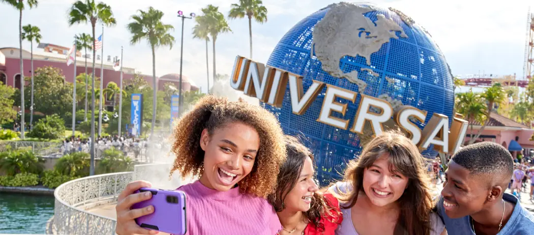A group of four friends takes a selfie in front of the iconic Universal globe, surrounded by palm trees and colorful signage
