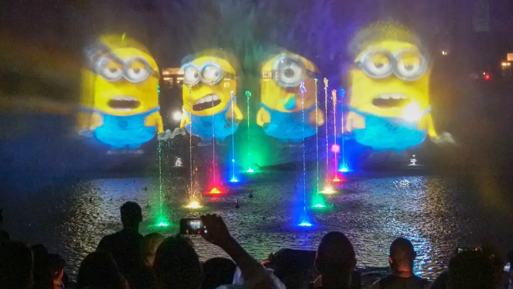 Vibrant, illuminated fountains dance in front of large Minion projections at night, captivating an audience from a waterfront view
