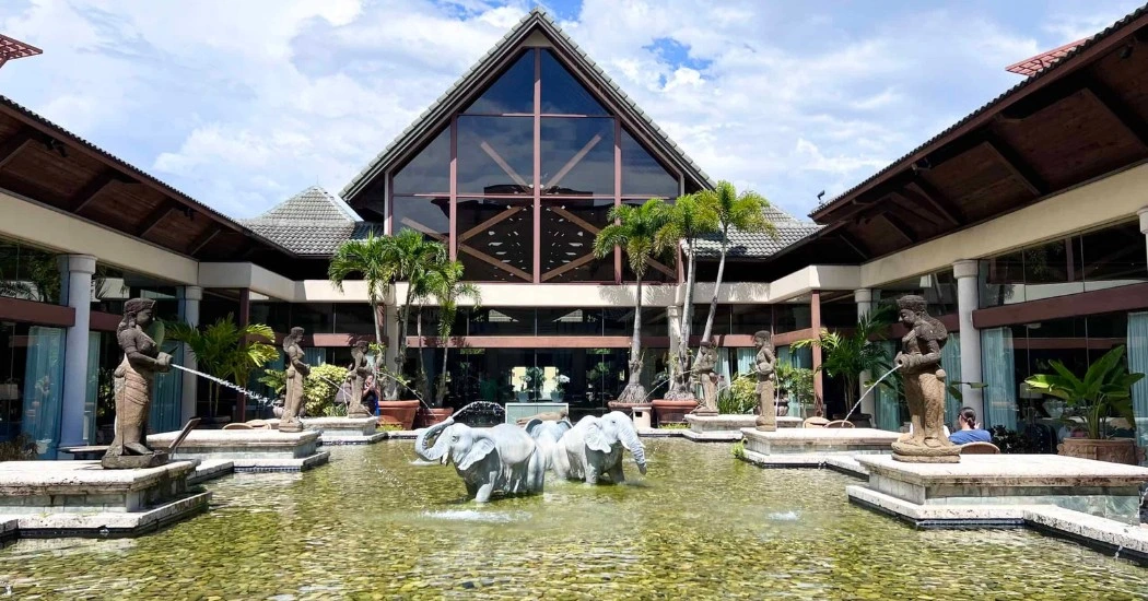 A luxurious entrance featuring clear water with elephant sculptures, ornate statues, lush greenery, and a large glass-fronted building