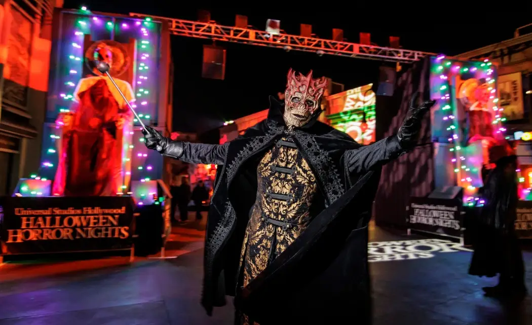 A skeletal figure in an ornate black cape poses dramatically at Halloween Horror Nights, surrounded by colorful lights and decorations