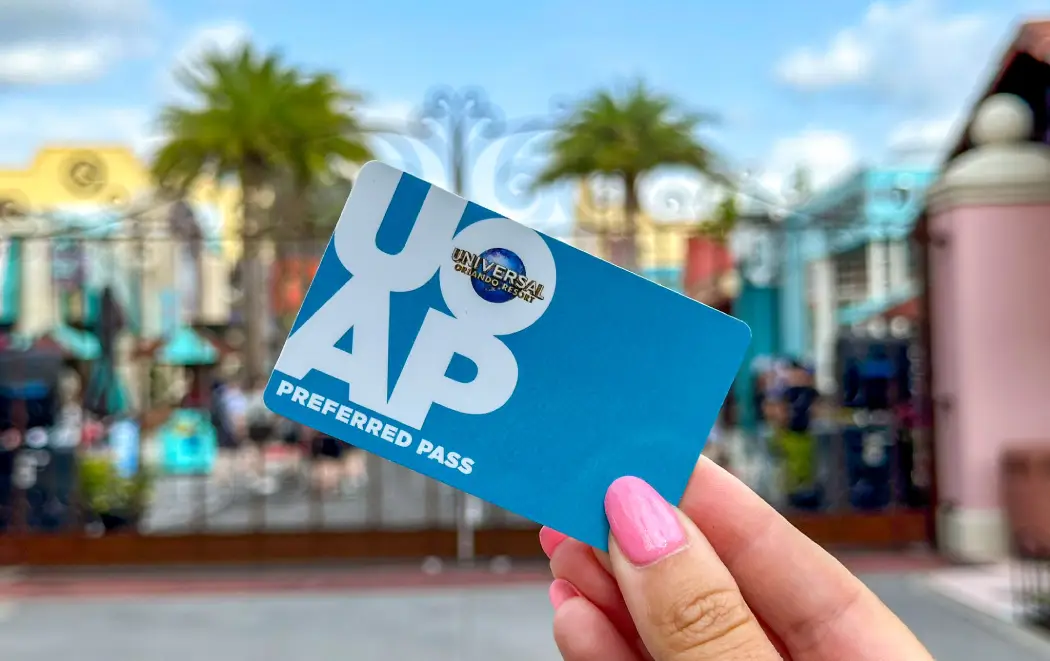 A hand holds a blue Universal Orlando Resort Preferred Pass in front of a vibrant amusement park background
