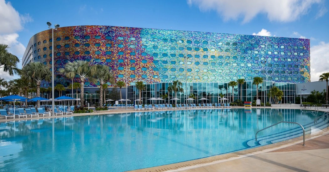 A colorful modern building with an oval window facade reflects in a pool surrounded by palm trees and lounge chairs